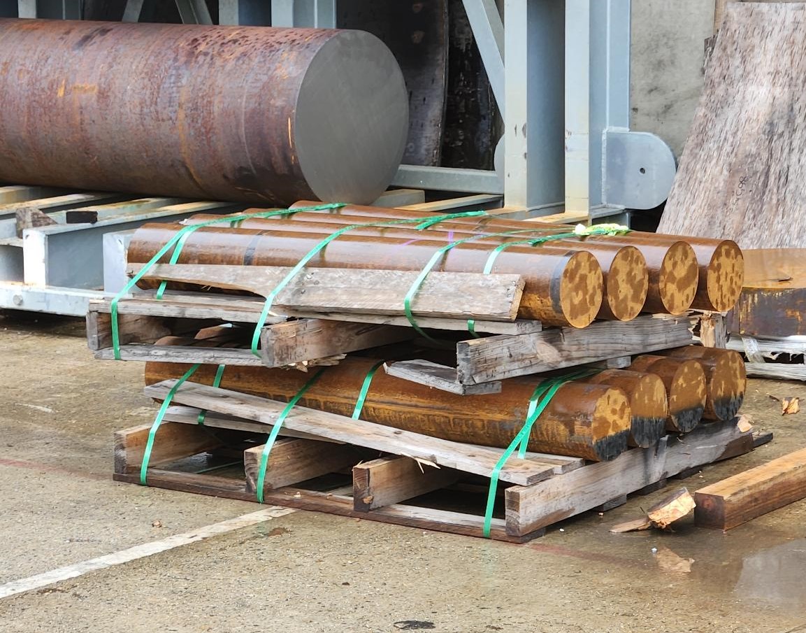 Cylindrical components in a yard, stored on stacked, broken timber pallets - showing signs of damage